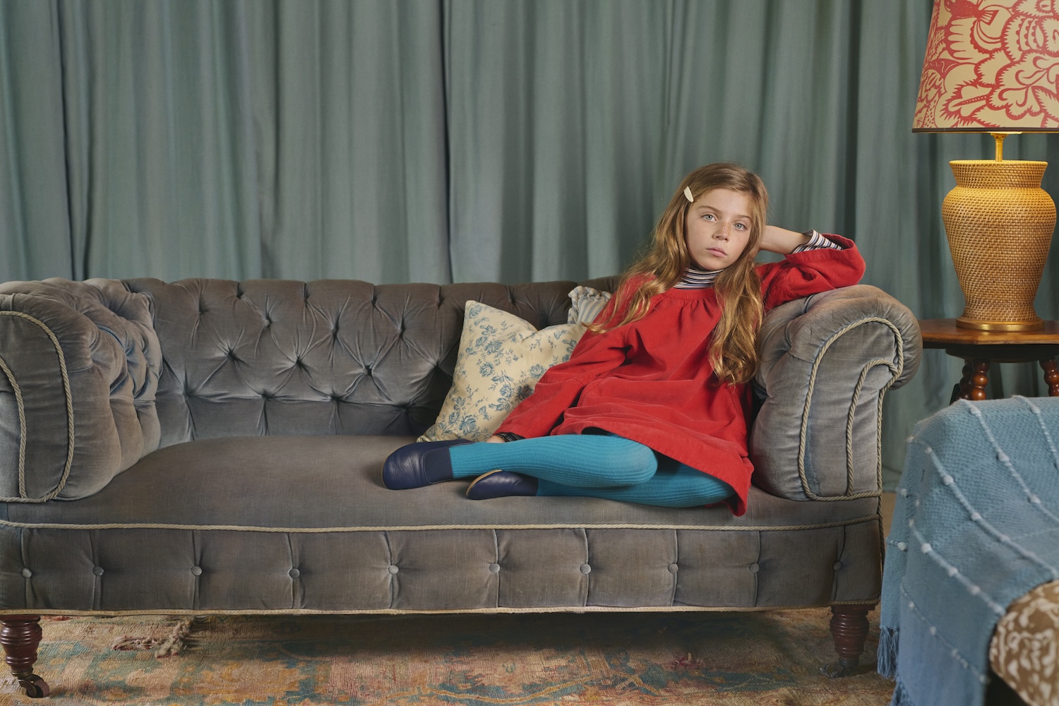 Child lounging on a vintage grey sofa in a red dress, turquoise tights and blue shoes from Caramel for Zara.