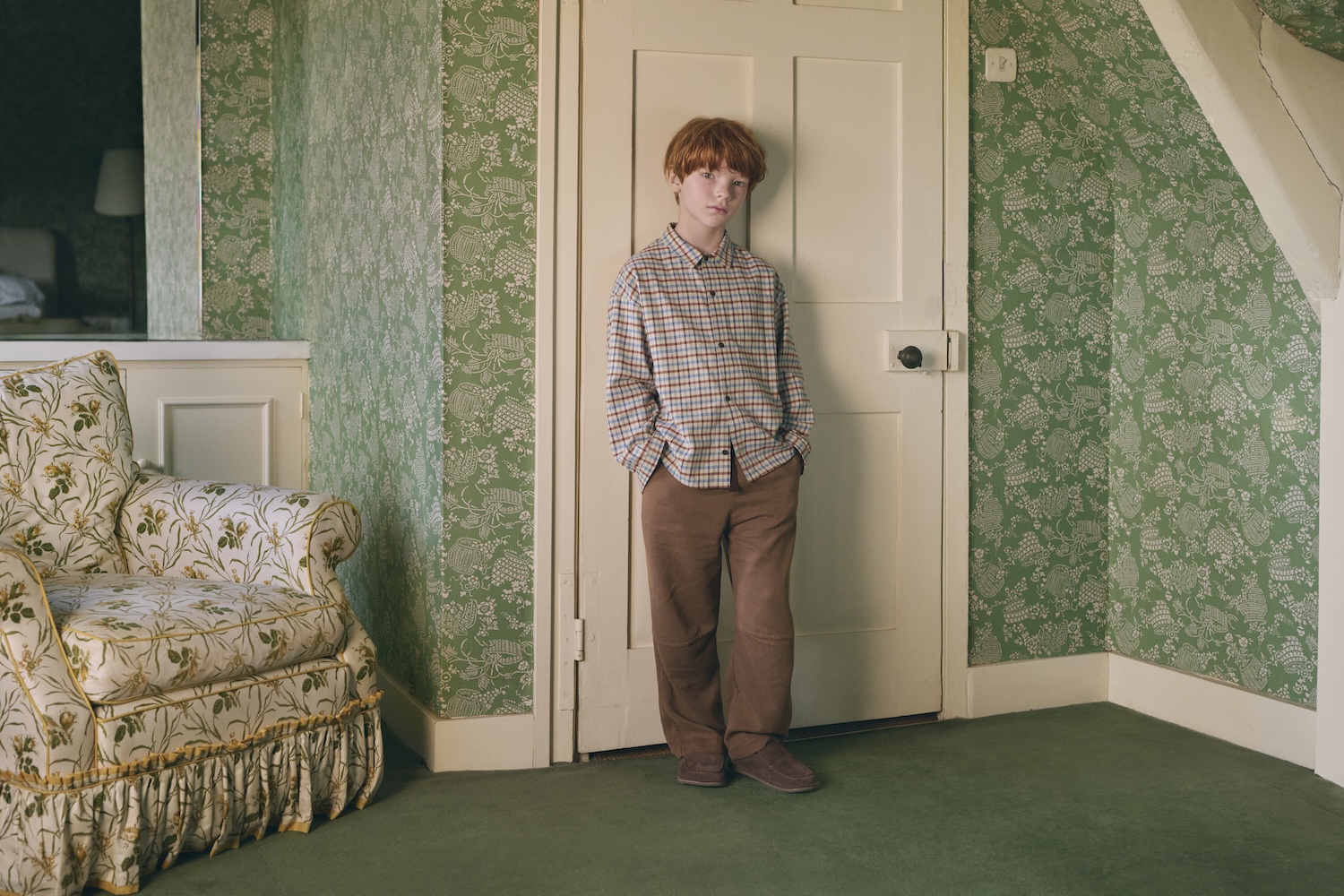 Child wearing a checked shirt, brown trousers and brown suede shoes, standing in a room with green floral wallpaper from Caramel for Zara.