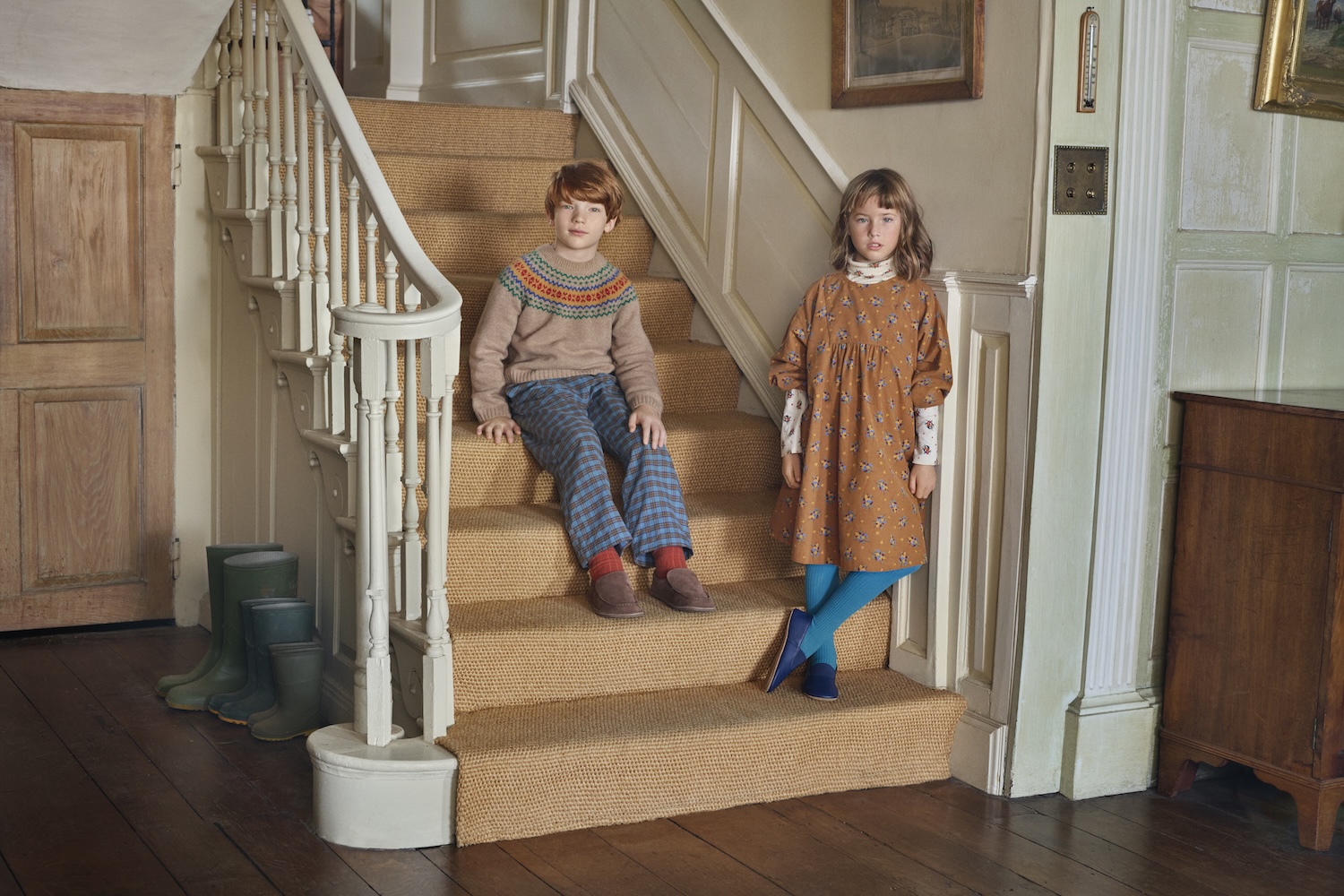 Two children wearing autumnal Caramel for Zara outfits, including a Fair Isle sweater with checked trousers and a floral dress with bright blue tights, sitting on a carpeted staircase.